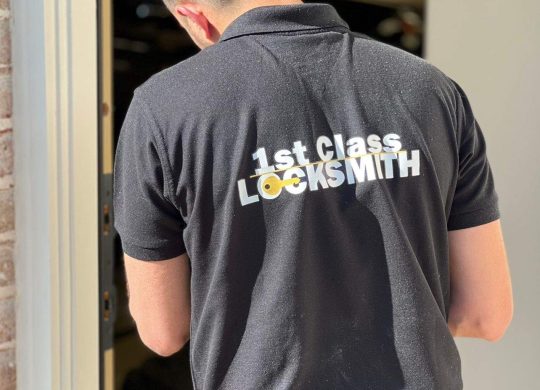 1st Class Locksmith technician working on a residential door lock, wearing a black uniform with yellow company branding in Dallas, TX